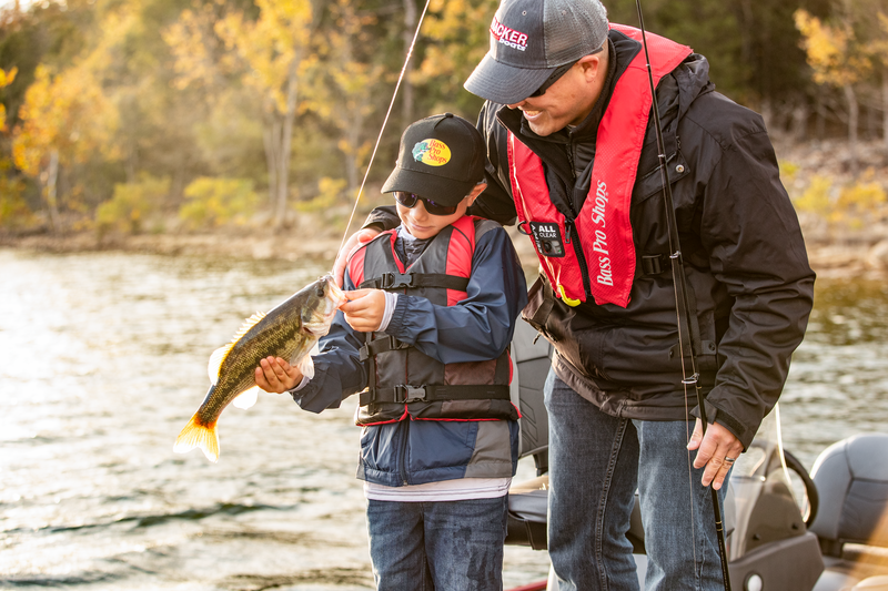 Father and son fishing together on a lake, proudly holding a largemouth bass while wearing Bass Pro Shops life jackets and outdoor apparel.