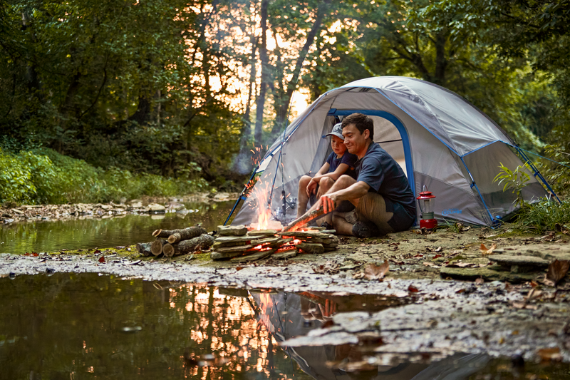 Couple enjoying a peaceful evening beside a creek with a tent and campfire, surrounded by nature and outdoor gear from Bass Pro Shops.