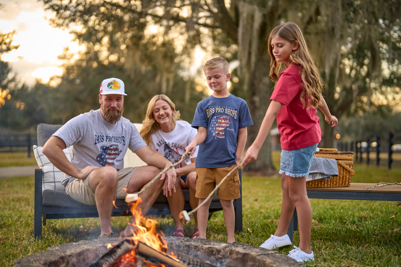 Family enjoying a campfire while wearing patriotic Bass Pro Shops Americana apparel, roasting marshmallows in a scenic outdoor setting.