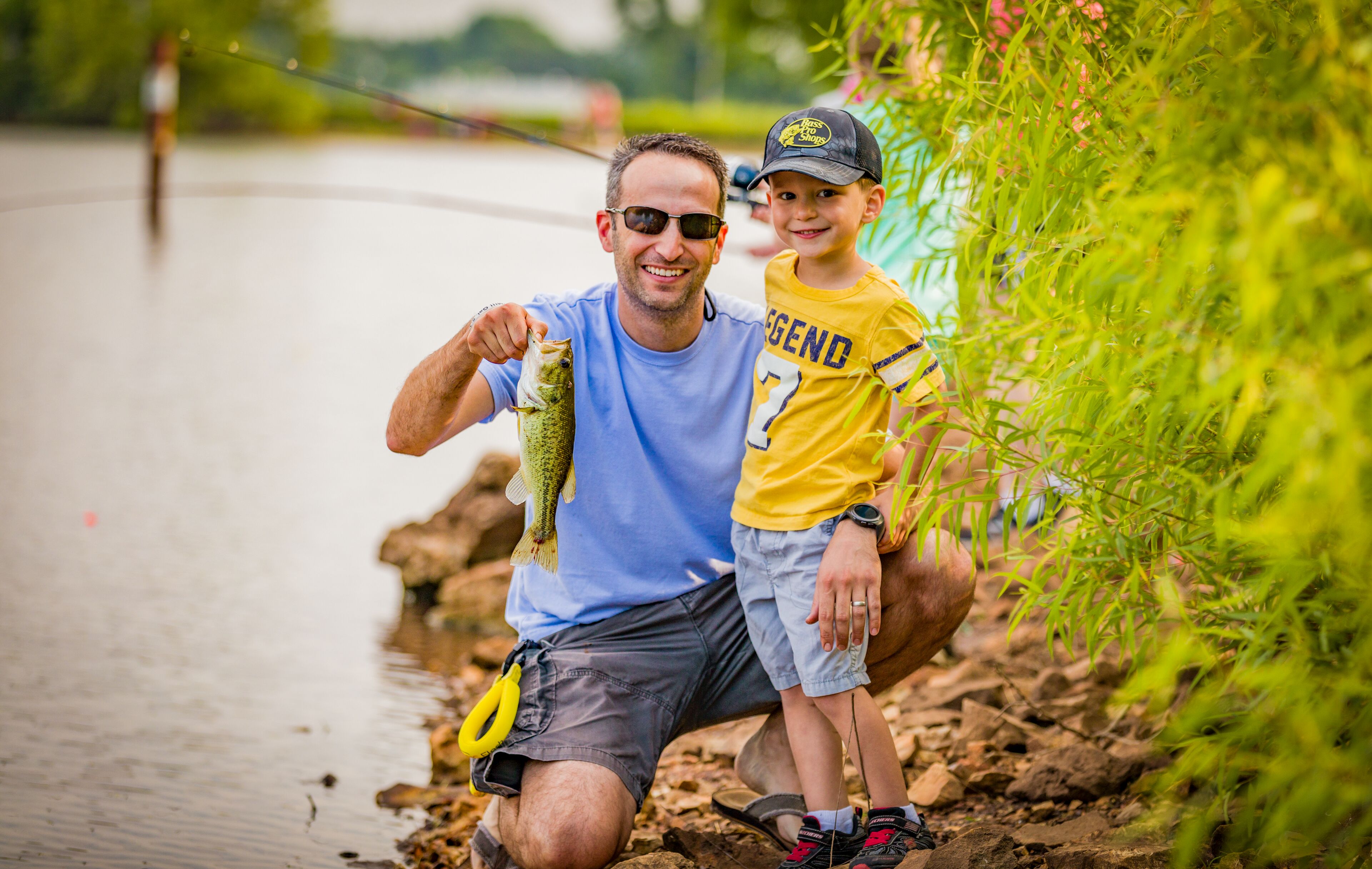 Father and son smiling by a lake as the father holds up a freshly caught fish.